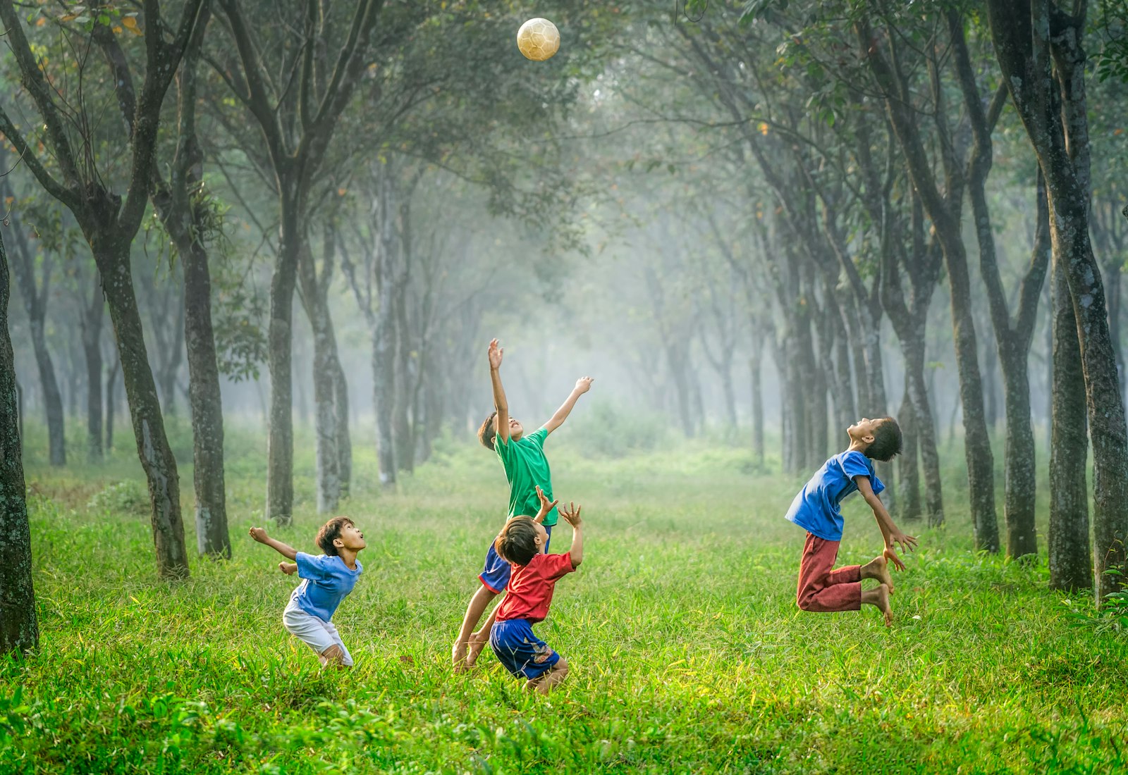 Four children laughing and leaping for a ball in a sunlit green park