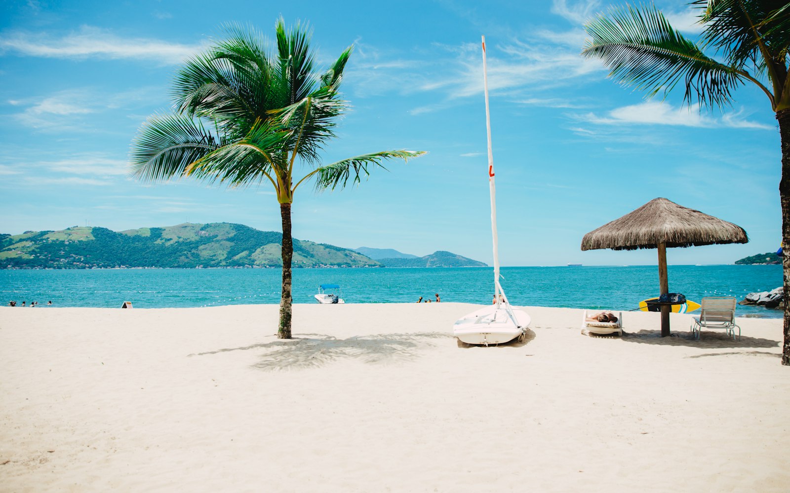 A sunlit tropical beach with palm trees and a small sailboat
