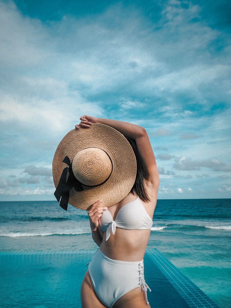 A model in a white bikini holding a straw hat by a turquoise pool