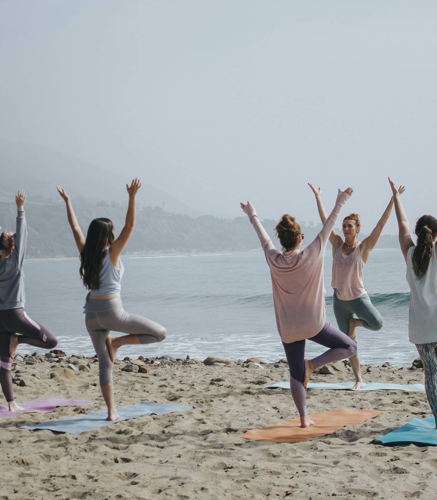 A small group flowing through tree pose on yoga mats beside the water at sunrise