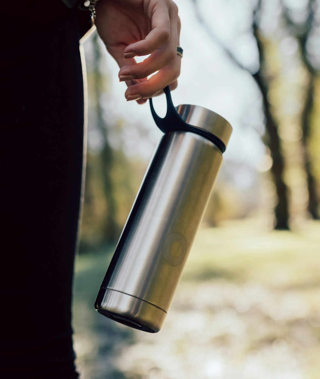 A person carrying a stainless steel Hale bottle by its handle through a sunlit park