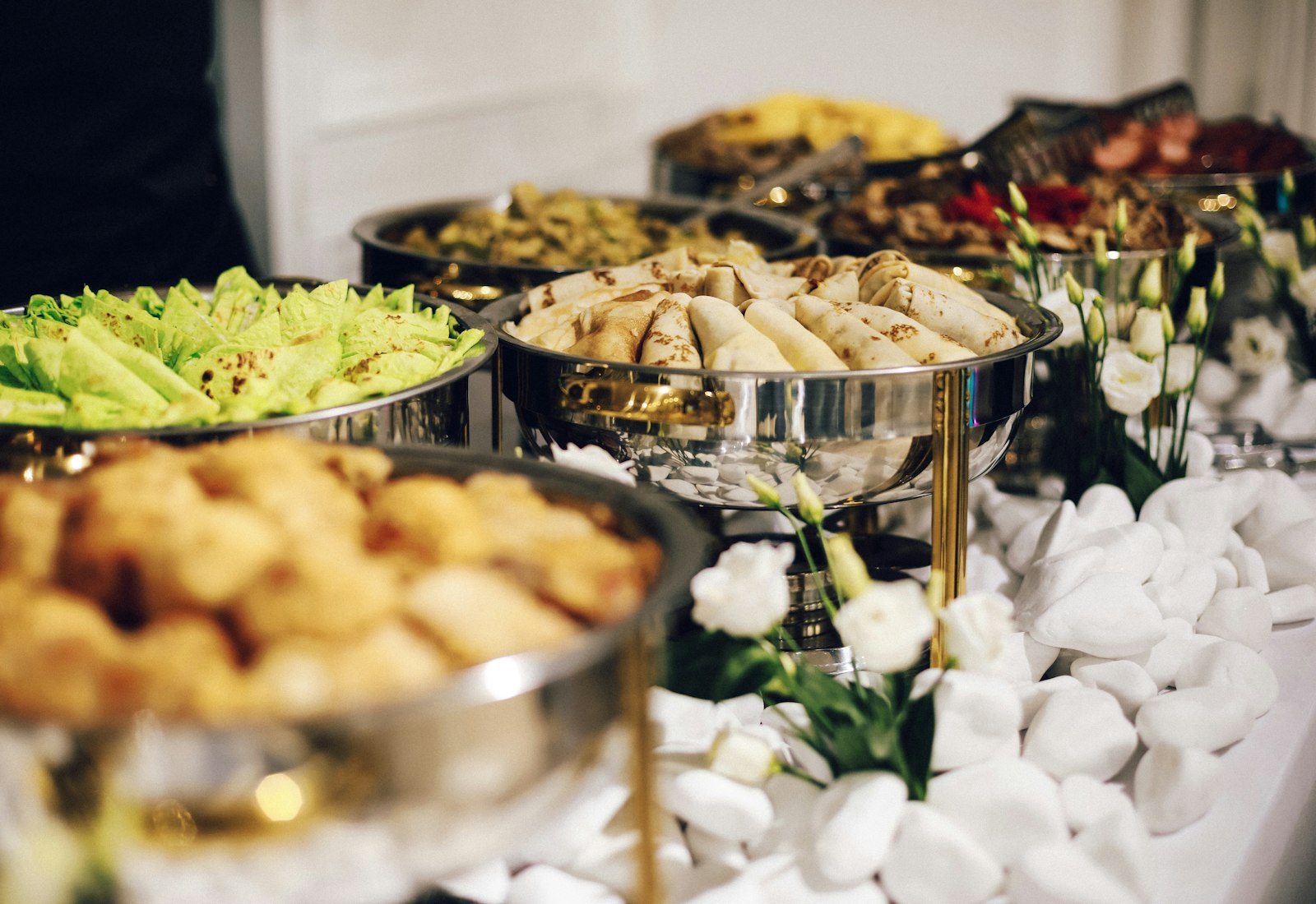 An elegant catered buffet of warm dishes in polished chafing bowls dressed with white roses.