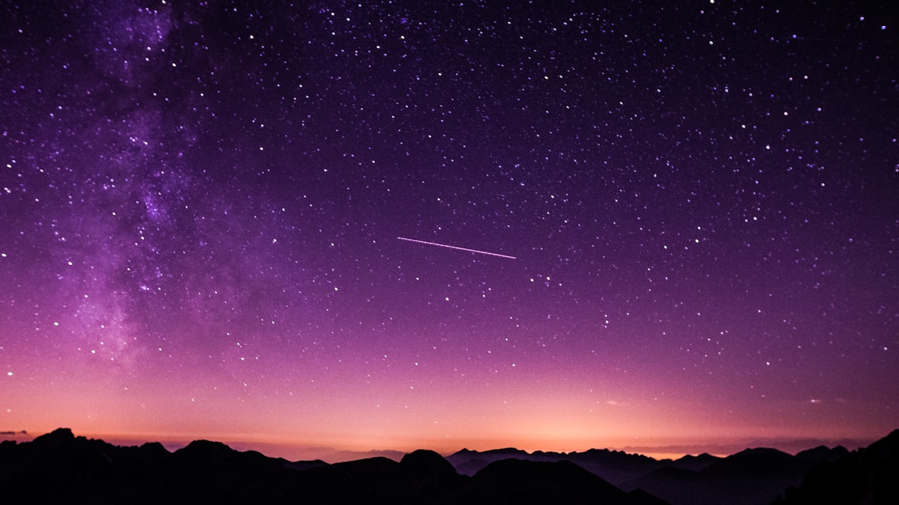 A violet star-filled sky with a shooting star arcing over silhouetted mountains.