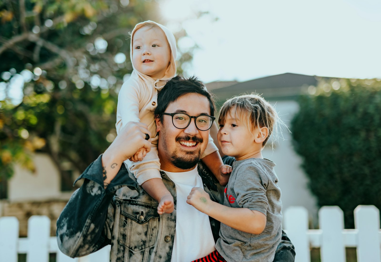 A smiling father in a denim jacket holding his two young children outside their home