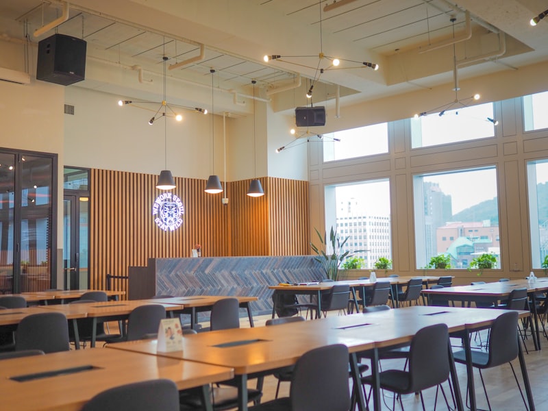 Long, sunlit communal work tables with pendant lamps and plants in the hot-desk area