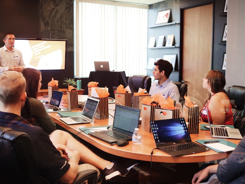 Meeting room with a large wall screen, presenter and members seated around the table with laptops