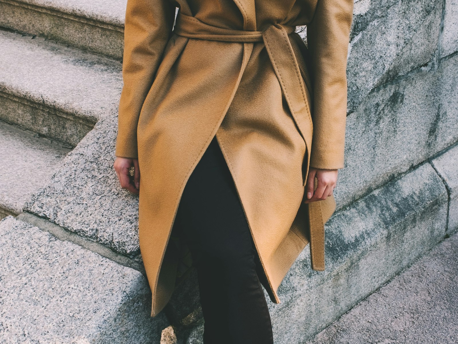 A model walking away from camera in a fluid camel wrap coat against a soft concrete backdrop