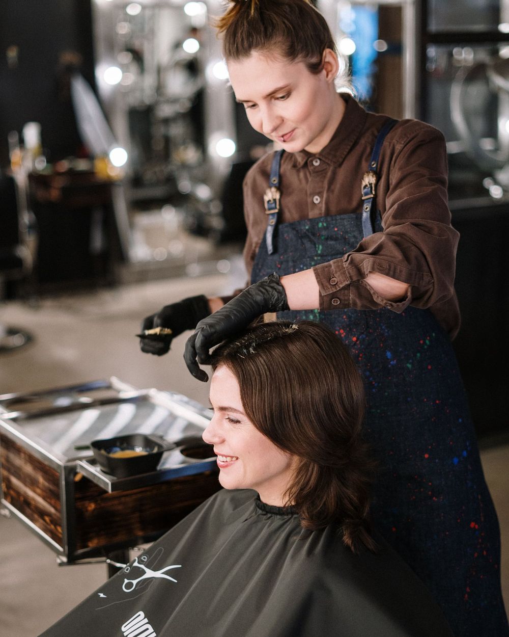 A colourist applies dye with gloved hands in a dark mirrored salon
